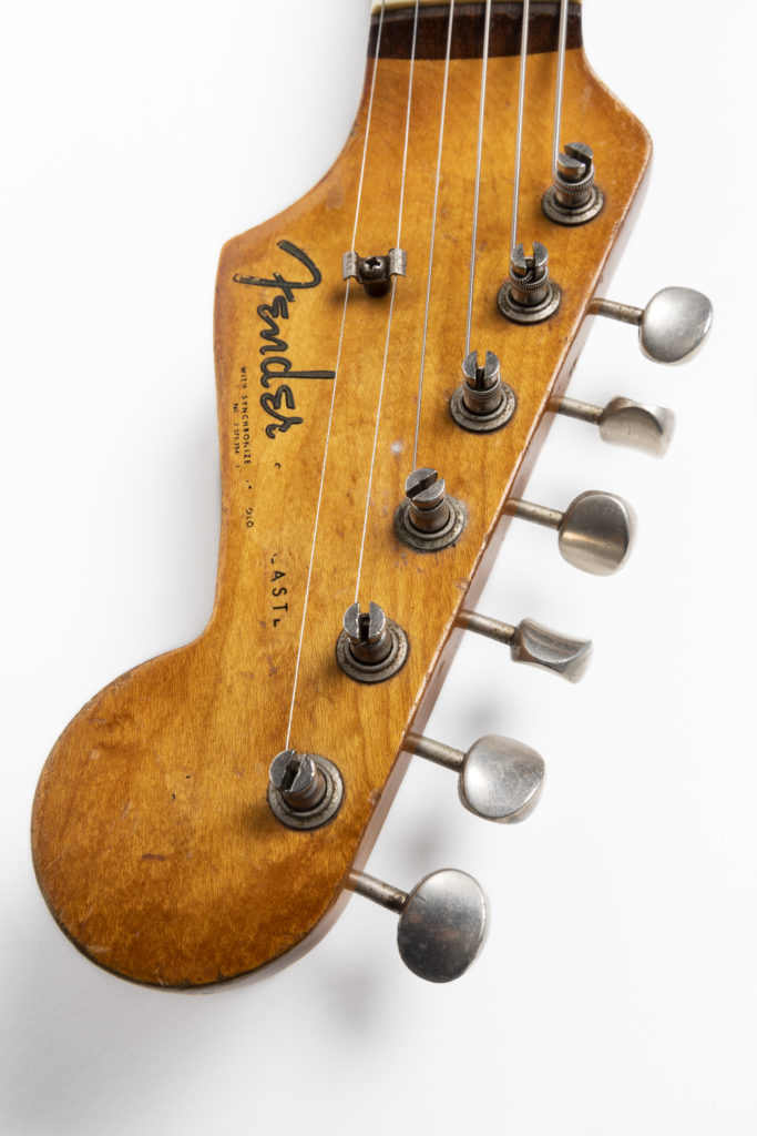 Detail of a Fender Stratocaster headstock. The headstock is made from a mid-coloured wood. There are six tuning pegs along the top, to which are attached six metal strings and a Fender logo along the bottom in the original 'spaghetti' style font.